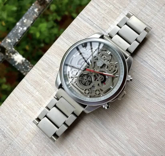 Tagheuer CR7 Chronograph Watch with grey stainless steel strap and transparent skeleton dial showing intricate gears and red-accented hands.